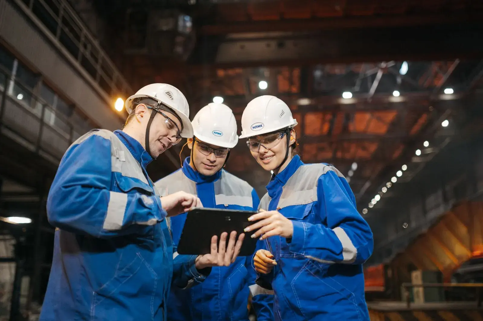 Three engineers in hard hats and blue overalls looking at a tablet on a factory floor.