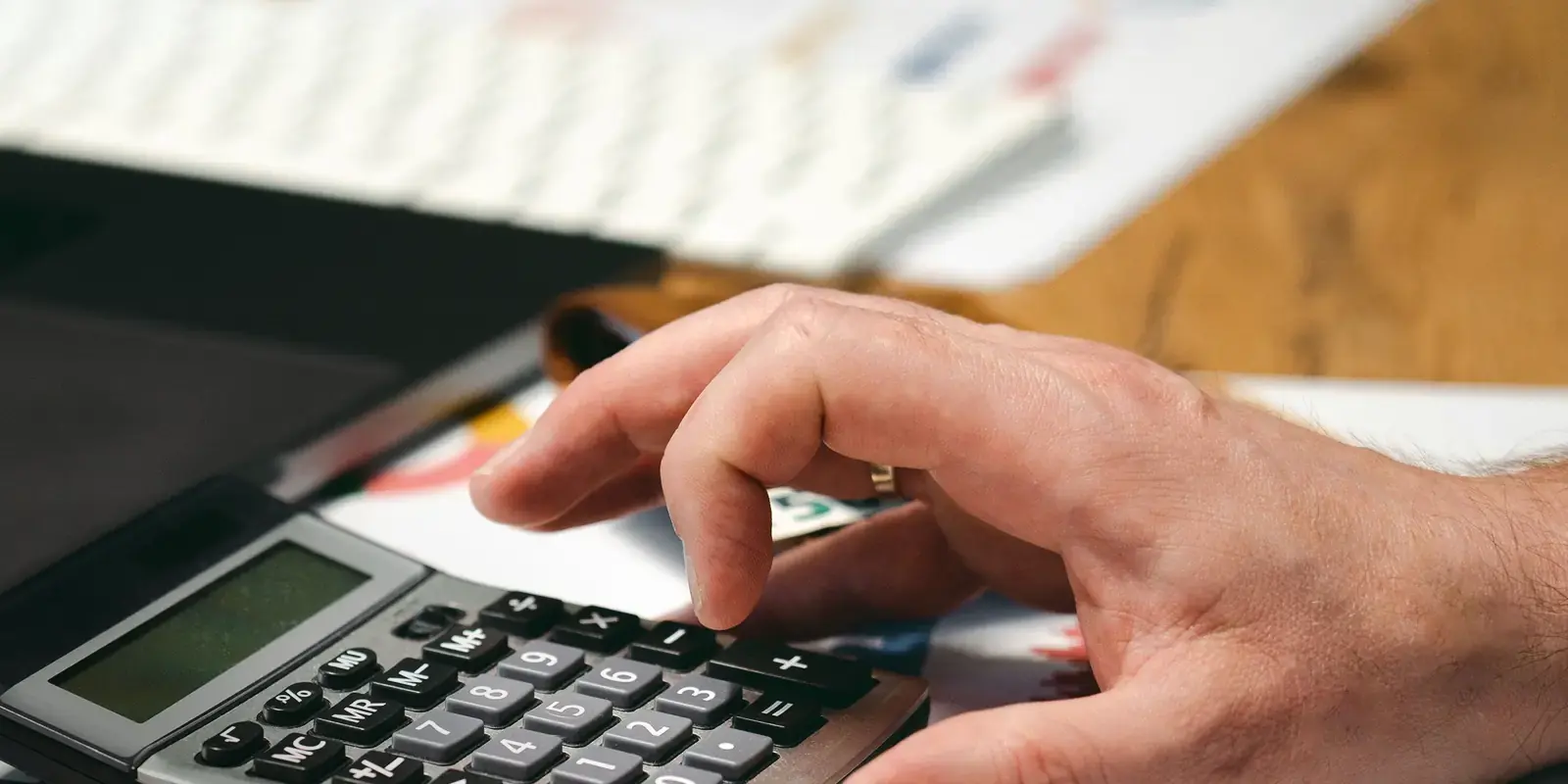 Hands working a calculator beside a laptop on a wooden desk.